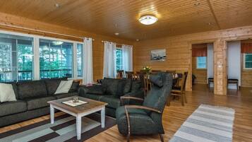 A spacious living room with a wooden ceiling, large windows, a sofa set, a coffee table, and a dining area.