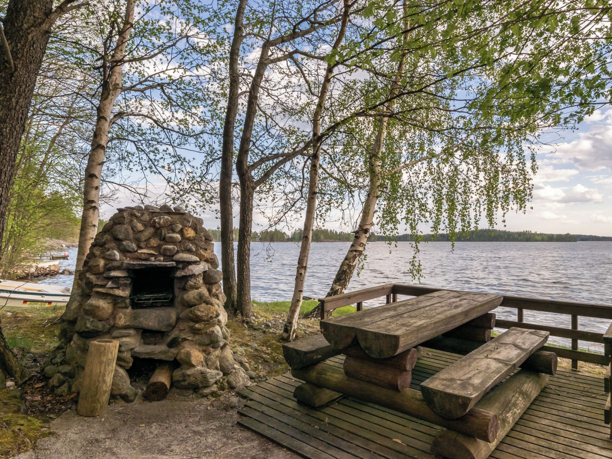 A stone-built outdoor cooking area with a wooden picnic table and benches, surrounded by trees and a lake.