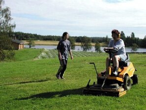 A person riding a lawn mower in a grassy field with a body of water and trees in the background. - Koivuniemi by Interhome (Sonkajärvi)
