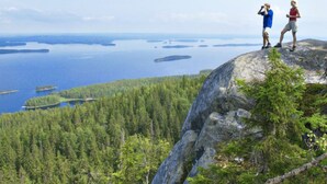 Kaksi ihmistä seisoo kallioisella kohdalla, josta avautuu näkymä metsäiselle alueelle ja vesistölle.