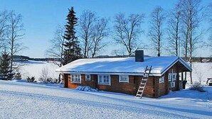 Lumihunnun peittämä puinen mökki, jonka seinää vasten nojaa tikapuut, ja taustalla kirkas, sininen taivas.