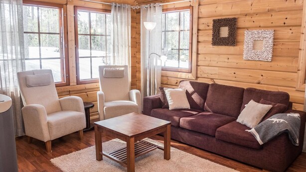 L’intérieur chaleureux d’un chalet, avec un revêtement mural en bois, un canapé, deux fauteuils, une petite table et de grandes fenêtres.