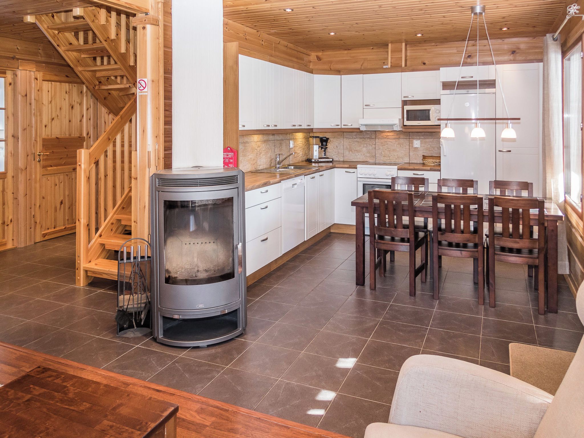 A modern kitchen with white cabinets, a built-in oven, and a stove. There is a dining area with wooden chairs and a table. A wood-burning stove is also visible.