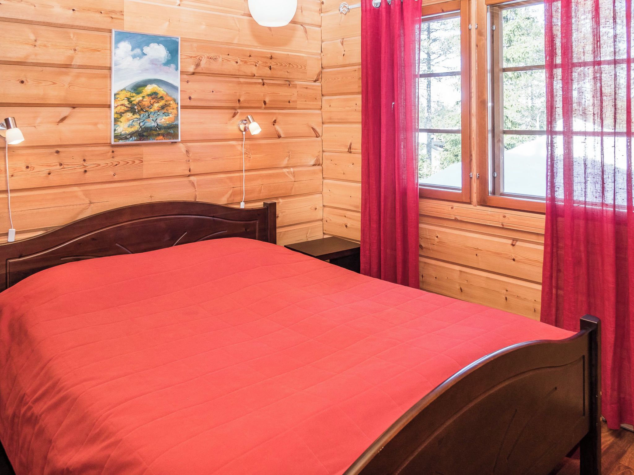 A wooden cabin room with a bed, a nightstand, a lamp, a painting, and a window with curtains.