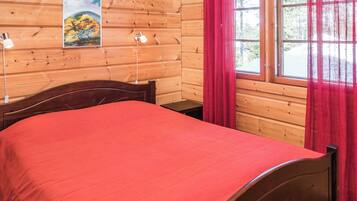 A wooden cabin room with a bed, a nightstand, a lamp, a painting, and a window with curtains.