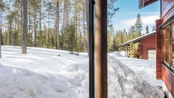 Uma cabana de madeira com um quintal coberto de neve e uma floresta ao fundo.