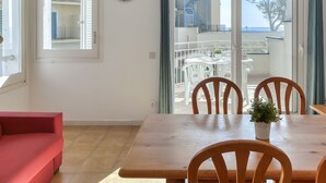 Ein Essbereich mit Holztisch und Stühlen, einem roten Sofa, einer Uhr und Blick auf das Meer durch eine Schiebetür.