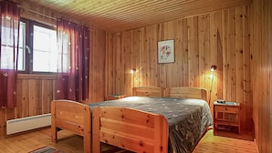 A wooden-paneled room with two wooden beds, a window with curtains, a framed picture on the wall, and a bedside table with a lamp. - Pajaranta by Interhome (Hartola)