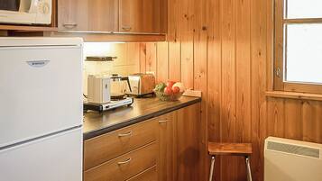 Een kleine keuken met houten kastjes, een witte koelkast, een broodrooster en een klein krukje.