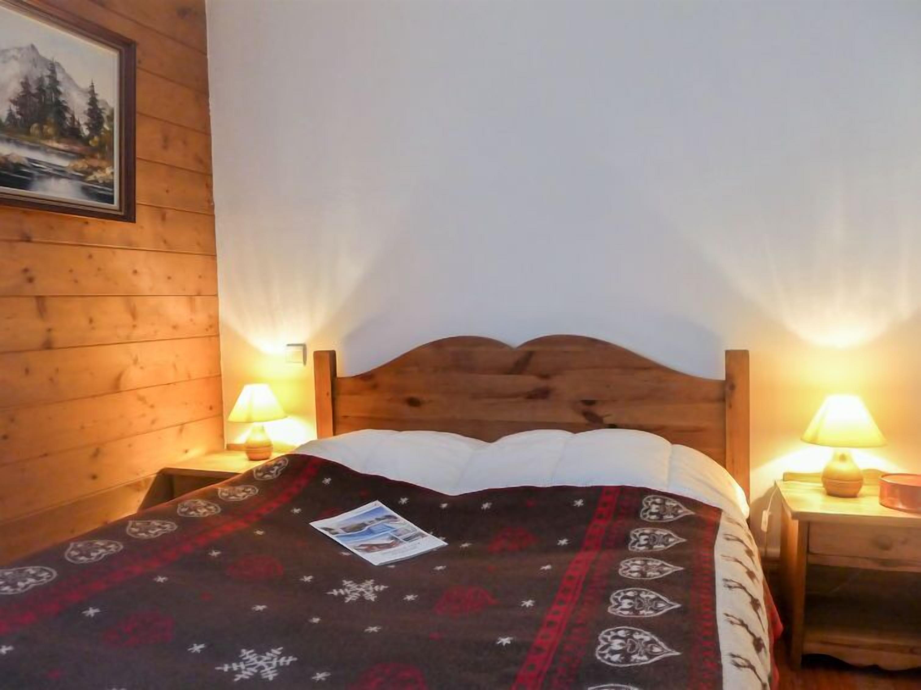 Una habitación de madera con una cama, dos lámparas de noche, un cuadro en la pared y una colcha estampada.