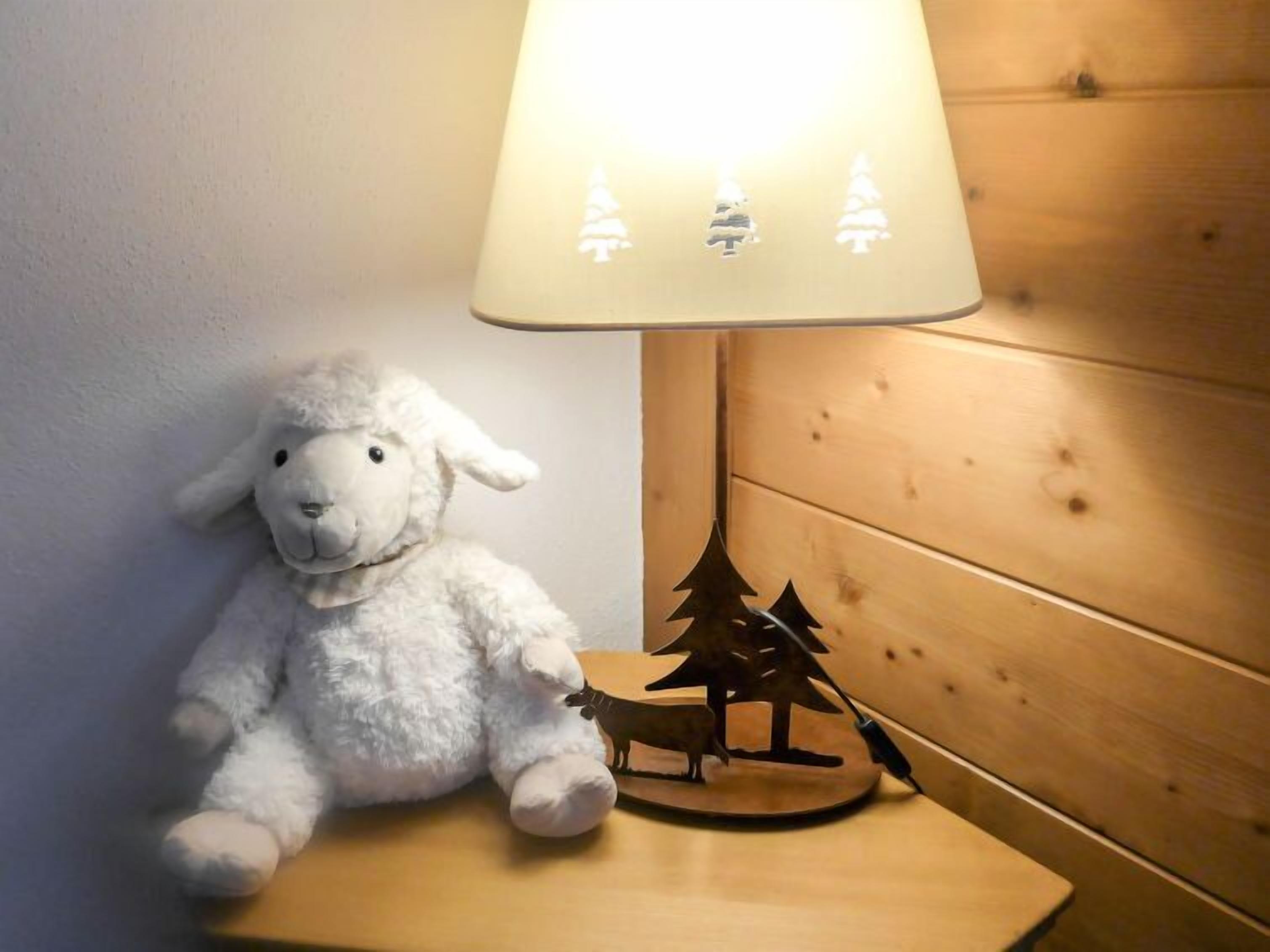Un mouton en peluche blanc est posé sur une table de chevet en bois, à côté d’une lampe dont l’abat-jour est décoré de motifs de sapin de Noël.