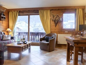 A cozy living room with a sofa, armchair, and coffee table, wooden walls, and large windows with curtains.
