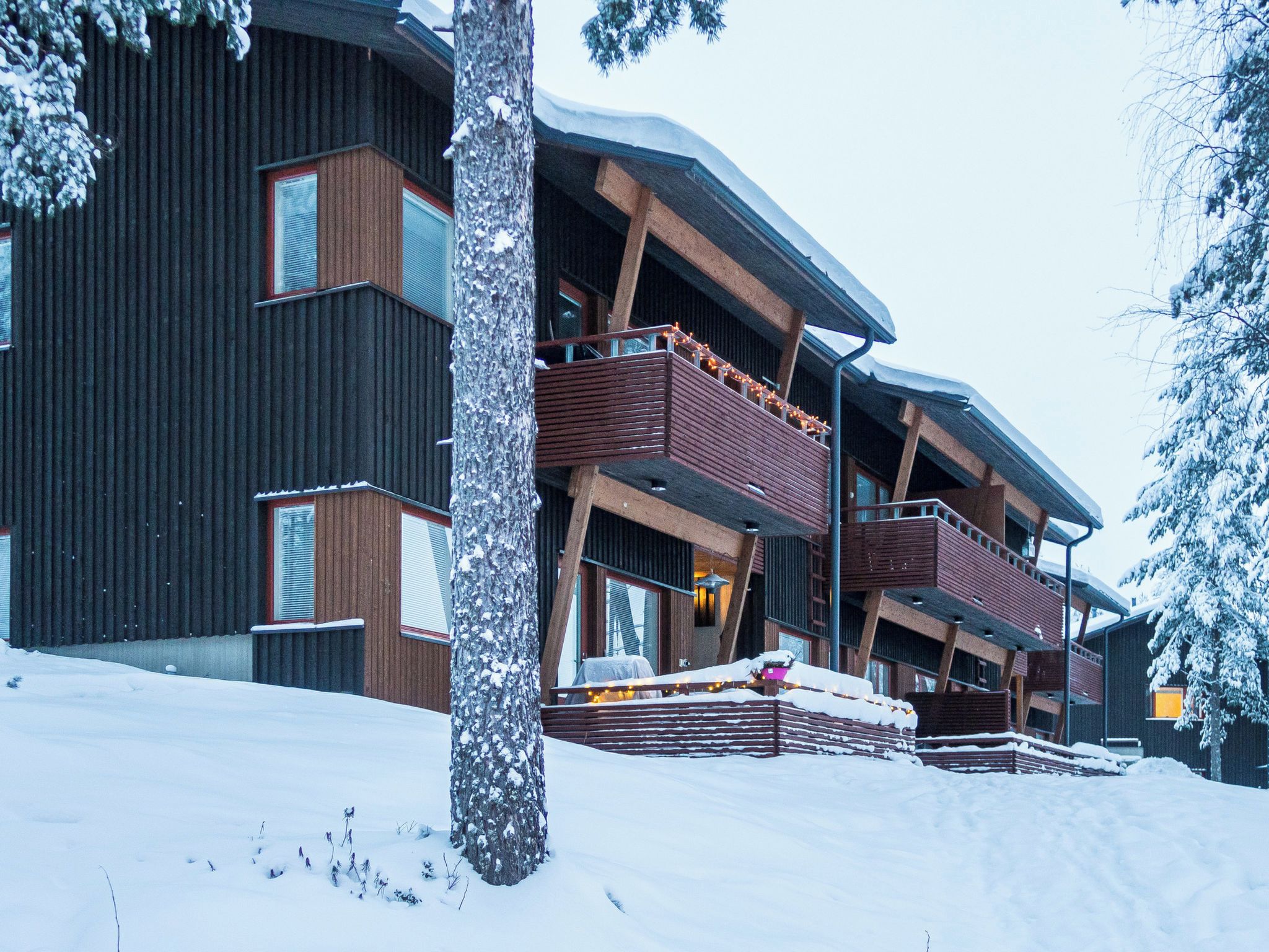 A multi-story building with balconies, a snow-covered ground, and a tree in the foreground.