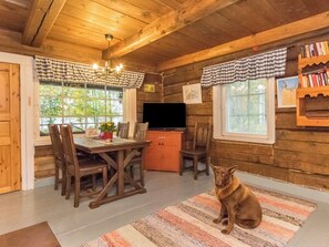 A cozy cabin interior with a wooden dining table, chairs, a television, and a dog sitting on a rug.