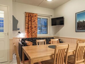 Tunnelmallinen huone, jossa on puinen ruokapöytä, tuoleja, kuviollisilla koristetyynyillä varustettu sohva, seinälle asennettu televisio ja ikkuna, jossa on sälekaihtimet.