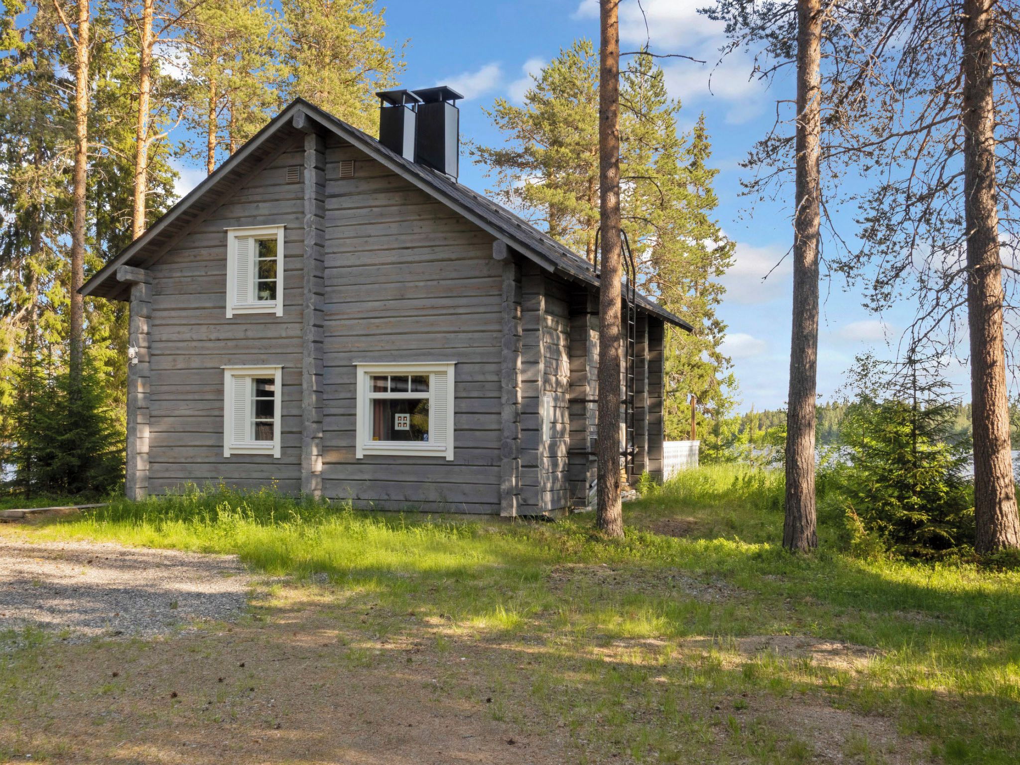 Kaksikerroksinen hirsihökkelö, jossa on savupiippu, ja sen ympärillä on puita ja kirkas taivas.