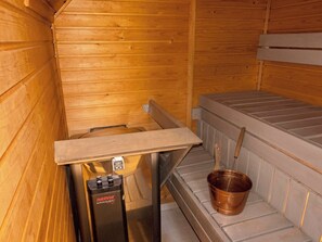 Un sauna con paredes de madera, un banco de metal, una pequeña mesa con un calentador y un cubo de cobre.