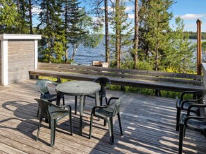 Una terraza de madera con una mesa redonda y sillas, con vista a un lago rodeado de árboles.