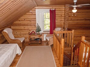 Acogedora habitación tipo cabaña de madera con techo inclinado, un pequeño árbol de Navidad, muebles de mimbre y una ventana con cortinas rojas.