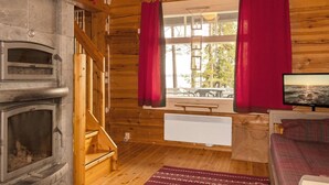 A wooden cabin interior with a stone fireplace, a wooden staircase, a window with red curtains, a television, and a view of the outdoors.