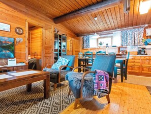 Ein gemütliches Cabin-Interieur mit Holzbalkendecke, einer Essecke mit blauen Stühlen, einer Couch mit gemusterten Kissen und einem Fernseher, der eine Berglandschaft zeigt.