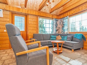 A cozy room with a wooden ceiling, a grey sofa, a wooden armchair, and a small wooden table with magazines. - Kinostupa 1a (2) by Interhome (Kuusamo)