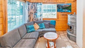 Interior acogedor de una cabaña con un sofá modular, una pequeña mesa redonda, una chimenea y grandes ventanales con persianas.