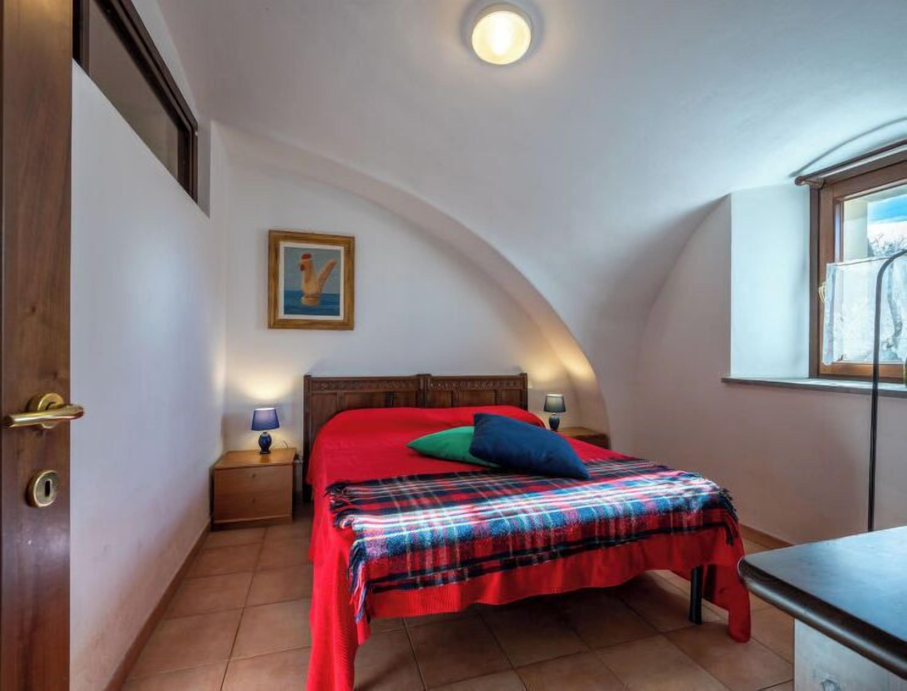 Une chambre à coucher avec un plafond incurvé, un lit recouvert d’une courtepointe rouge et d’une couverture à carreaux, une table de chevet avec une lampe, et un tableau encadré accroché au mur.