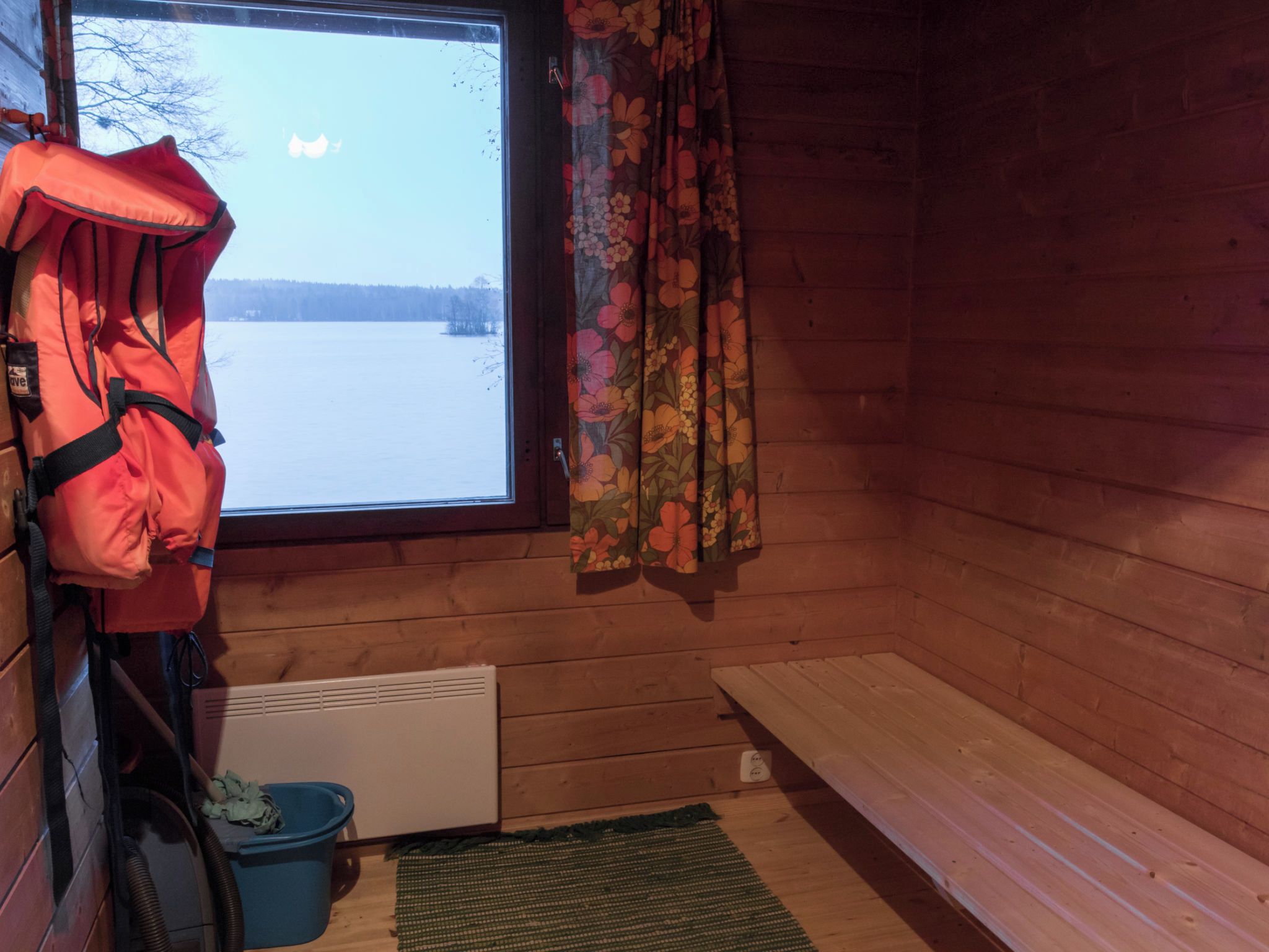 A cabin interior with a window, a bench, a heater, and a blue bucket.