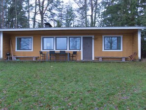 Eine eingeschossige Holzhütte mit Veranda, vier Fenstern und einem kleinen Tisch mit Stühlen.