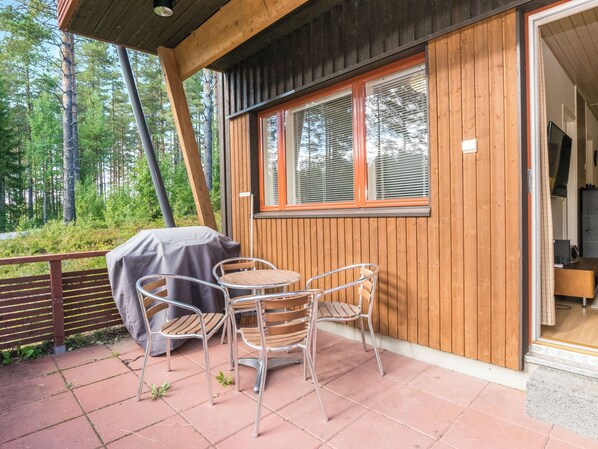 A wooden cabin with a patio area featuring a table and chairs, a covered grill, and a view of the surrounding forest.