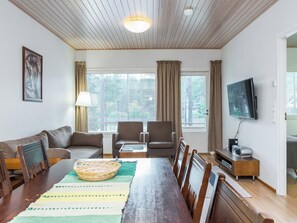 Un salon moderne avec un plafond en bois, un canapé, deux fauteuils, une table à manger avec des chaises, un téléviseur et une fenêtre avec des stores.