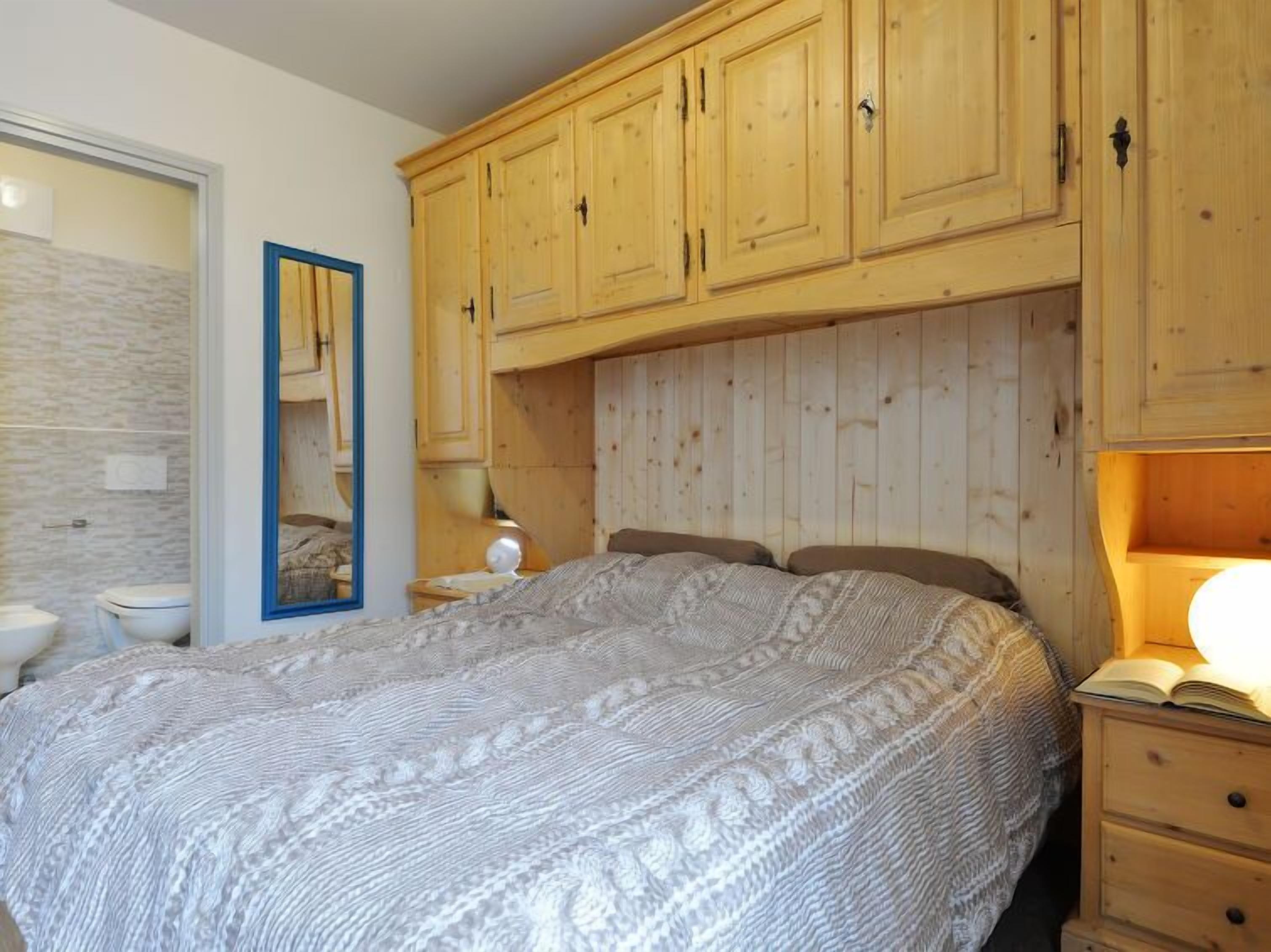 A bedroom with a large bed, wooden cabinets, a mirror, and a bedside table with a lamp.