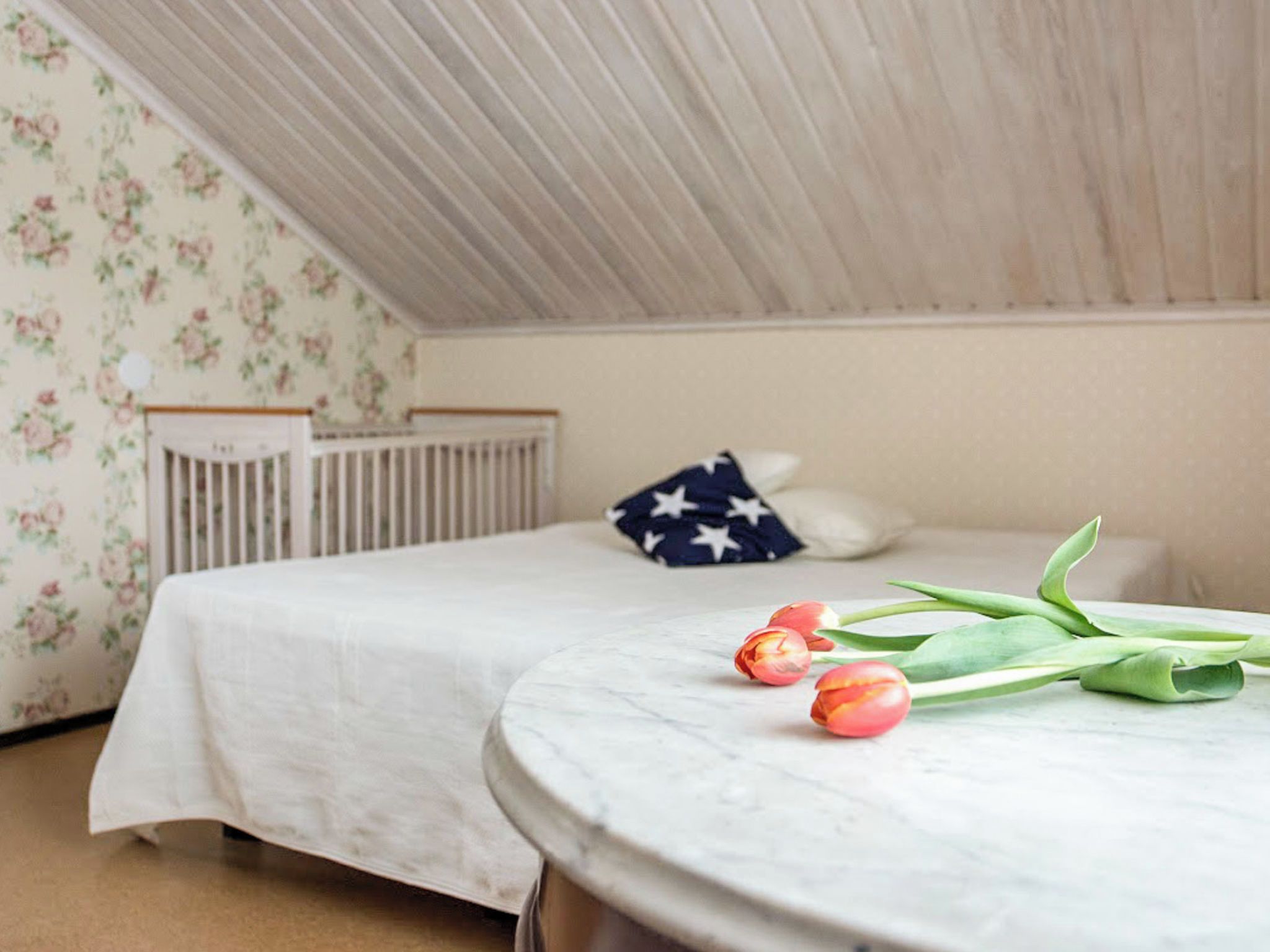 A bedroom with a bed, a crib, a round table with tulips, and a wall with floral wallpaper.