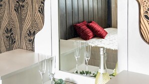 Ein weißer Waschtisch mit Spiegel, drei leere Weingläser, eine Champagnerflasche und ein rotes Kissen auf einem Bett mit weißer Bettdecke.
