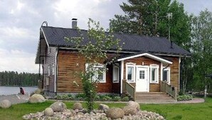 Eine Holzhütte mit Veranda, ein kleiner Garten mit Steinen und ein See im Hintergrund.