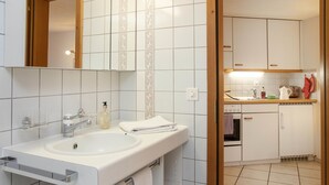 Un baño moderno con lavamanos, espejo y toallero. Junto al baño, una cocina compacta con estufa, horno y encimera.