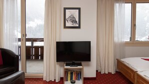 Una habitación de hotel con una cama, un televisor sobre un mueble, una estantería con libros y una ventana con cortinas.
