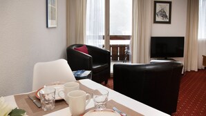 Habitación de hotel con una mesa de comedor preparada para dos personas, un sofá, un televisor y una ventana con cortinas.