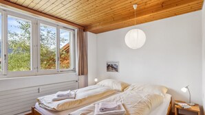 Ein Schlafzimmer mit zwei Betten, Holzboden, einem Fenster mit Vorhängen, einer Pendelleuchte und einem kleinen Nachttisch mit einer Lampe.