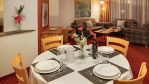 Uma sala de jantar com uma mesa redonda posta para quatro pessoas, rodeada por cadeiras de madeira. Há um vaso com flores sobre a mesa e também se vê uma garrafa de vinho. O ambiente é acolhedor, com um sofá e uma pequena mesa ao fundo.