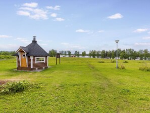 A small wooden cabin with a chimney, a lamp post, and a grassy field with trees in the background.