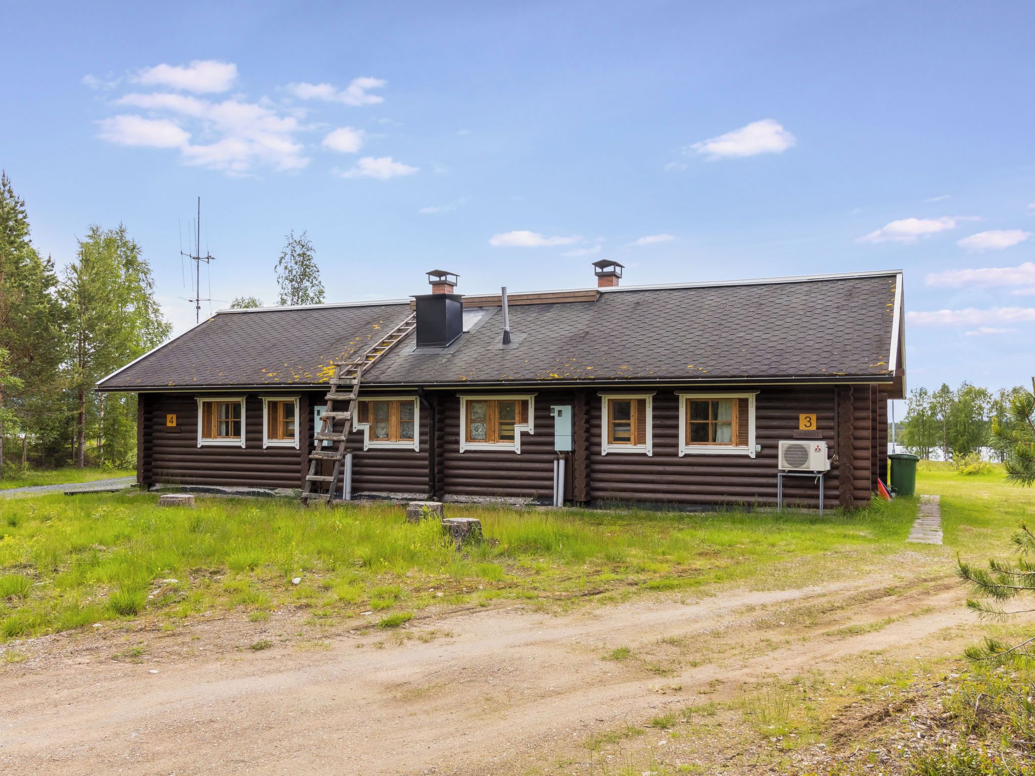 Une cabane en rondins, avec un chemin de terre menant à l’entrée, une échelle et un petit porche.