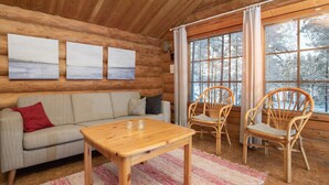 Un accogliente interno di una baita con soffitto in legno, un divano, due poltrone in vimini, un tavolo in legno e una grande finestra con vista innevata.