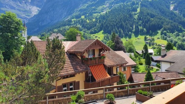 Un chalet doté d’un balcon, entouré d’une végétation luxuriante et avec des montagnes en toile de fond.