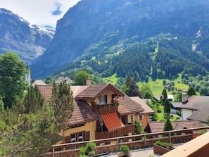 A chalet with a balcony, surrounded by lush greenery and mountains in the background.
