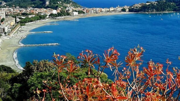 A coastal town with a mountainous backdrop, a clear blue sea, and a pier extending into the water.
