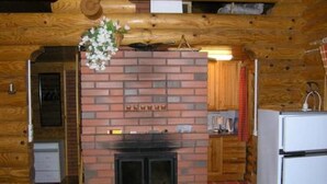 A log cabin interior with a brick fireplace, wooden walls, a kitchen area with a refrigerator, and a hanging flower arrangement. - Kettupirtti by Interhome (Juva)