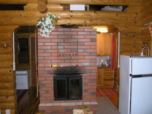 A log cabin interior with a brick fireplace, wooden walls, a kitchen area with a refrigerator, and a hanging flower arrangement. - Kettupirtti by Interhome (Juva)