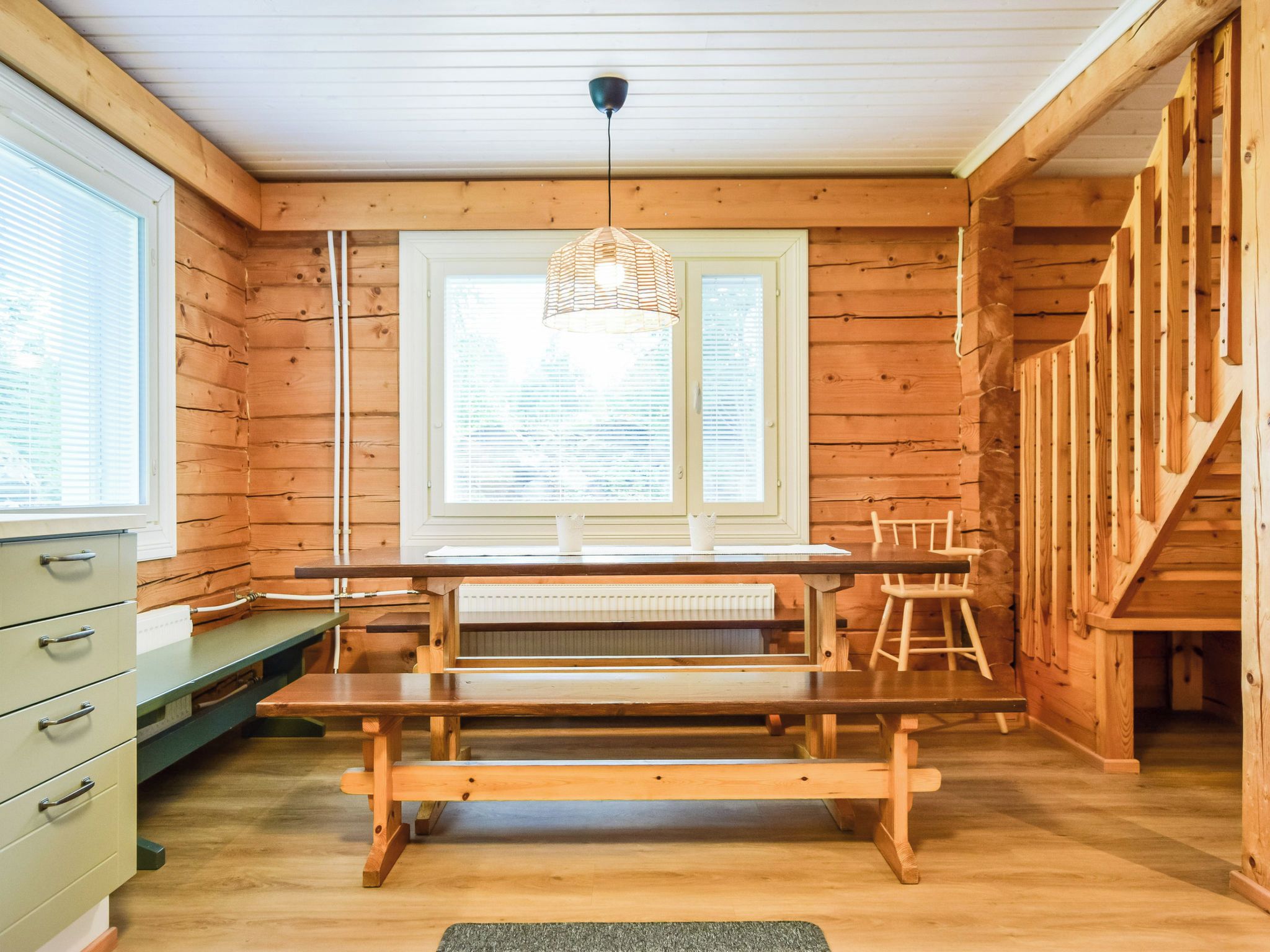 Interior de madera con una mesa de comedor, bancos, una ventana con persianas, una lámpara colgante y una escalera.
